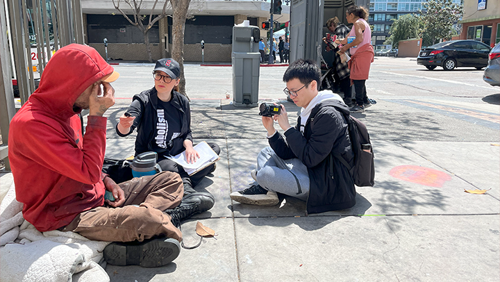 Woman interviews  an unhoused community member downtown while male student  vidoe tapes the interview