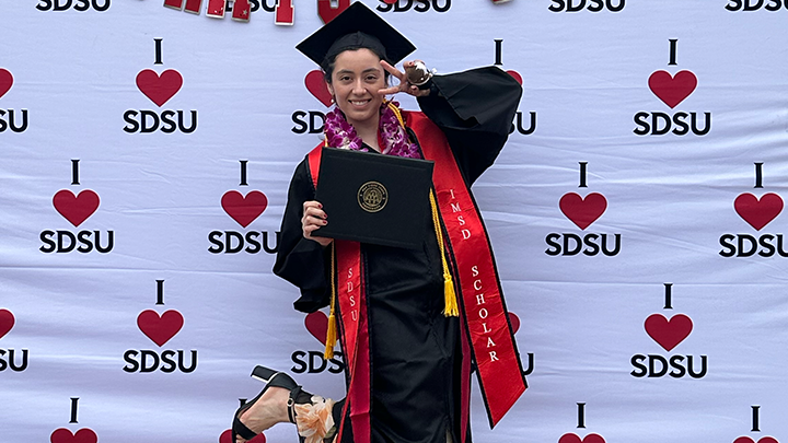 graduate in cap and gown stands poses in front of I heart SDSU