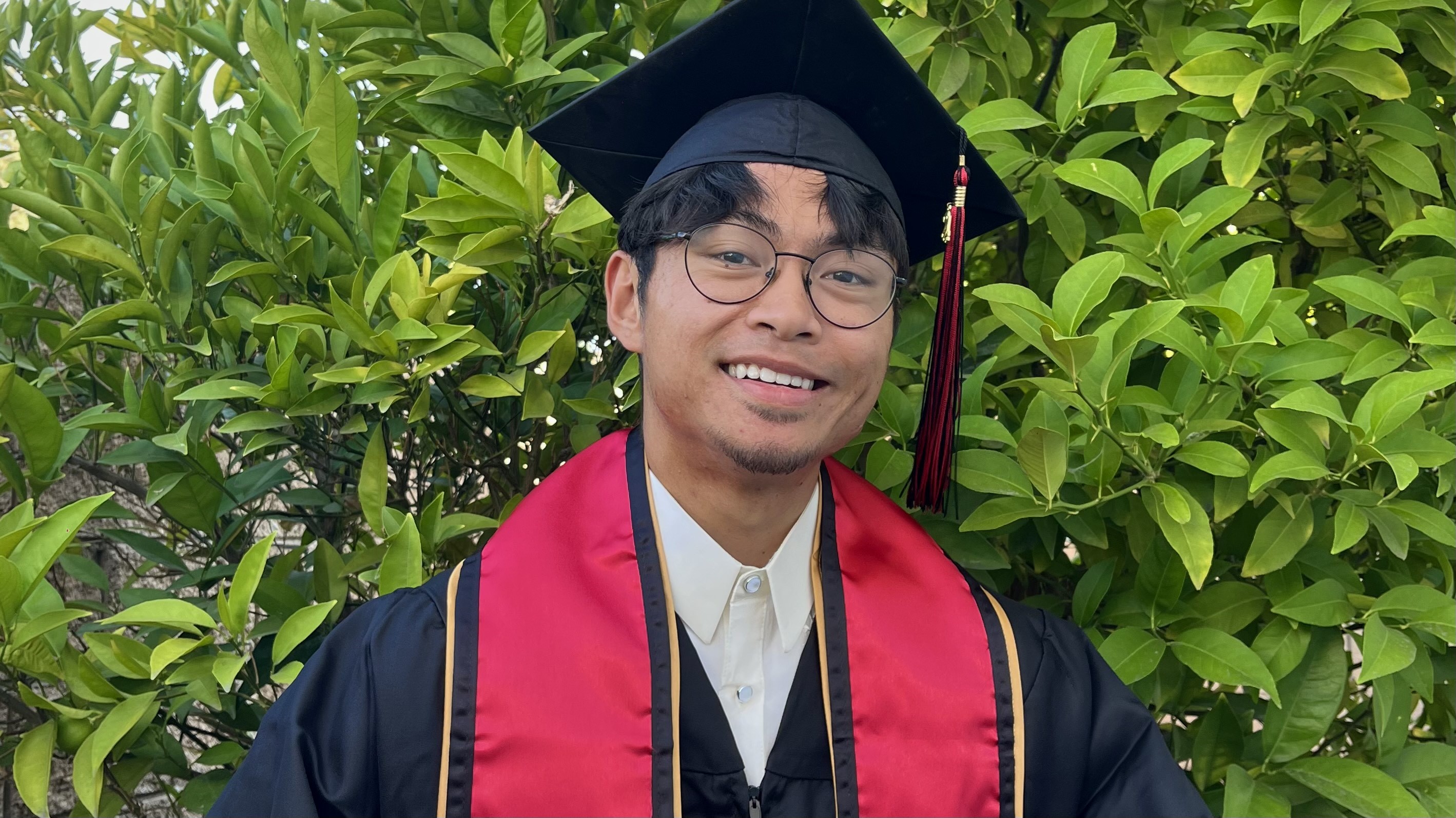student with glasses  in cap and gown smiles