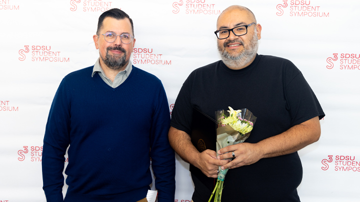 two smiling men pose at awards, one holds bouquet of flowers